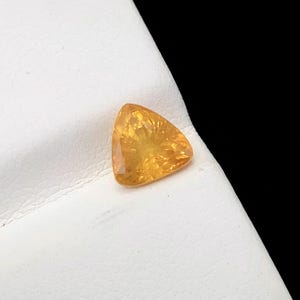 May include: A triangular, faceted gemstone in a warm, golden-yellow colour. The gem is set against a white surface with a black background, highlighting its clarity and brilliance. The gemstone's cut showcases its internal structure and light play.