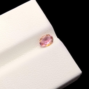 Puede incluir: Una piedra preciosa de corte ovalado con un degradado de tonos rosa suave y amarillo cálido. La piedra muestra una superficie facetada que refleja la luz, exhibiendo una claridad translúcida y un acabado pulido sobre una base blanca.