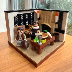 May include: A Lego diorama depicting a medieval kitchen scene. Two minifigures stand in a room with a fireplace, table, and barrel. The scene includes candles, a bowl, and a bottle. The diorama is set on a wooden surface.