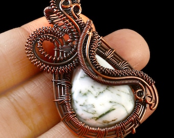Peut inclure: Pendentif artisanal en fil de cuivre, orné d'une pierre blanche en forme de goutte d'eau avec des veines vertes. La pierre est finement enveloppée de fil de cuivre, créant un design unique et artistique. Le pendentif est tenu dans une main sur fond noir.
