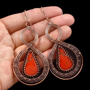 May include: Copper wire earrings with a teardrop-shaped red stone inlay. The earrings have a woven wire design around the stone and a looped top. The background is black.