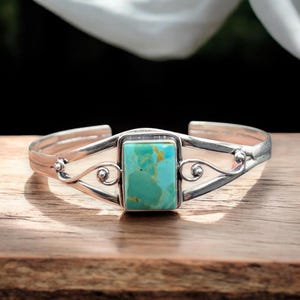 May include: A silver cuff bracelet featuring a rectangular turquoise stone set in the centre. The metal band displays intricate swirling wire details on both sides of the stone, with a polished finish and an open back design for easy adjustment.