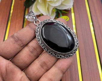 May include: An oval pendant featuring a large, smooth, polished black stone set in a decorative silver frame with intricate filigree scrollwork. The pendant includes a silver bail at the top for attachment to a chain or cord necklace.