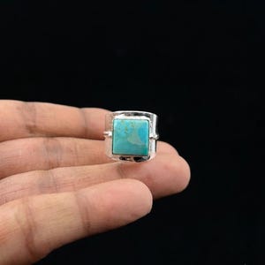 Puede incluir: Un anillo de plata con una piedra turquesa de corte cuadrado y vetas naturales. La piedra está engastada en un bisel de metal liso, montada sobre una banda de plata ancha y pulida que rodea el dedo con un acabado metálico limpio.