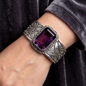 Peut inclure: Un large bracelet manchette en argent orné d'un motif de volutes complexe et d'une grande pierre précieuse rectangulaire à facettes violette au centre. Le métal présente une finition polie avec des zones sombres pour souligner le design.