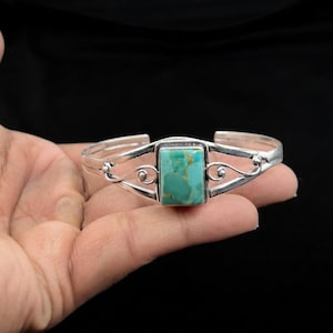 Könnte beinhalten: Ein silbernes Armband mit einem rechteckigen Türkisstein in der Mitte. Das Metallband weist auf beiden Seiten des Steins dekorative, geschwungene Drahtdetails auf, die ein durchbrochenes Design bilden, das sich zu den Enden hin verjüngt.