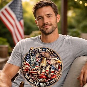 K&ouml;nnte beinhalten: Ein graues T-Shirt mit einem patriotischen Design. Das Design enth&auml;lt den Text "1776-2026 250 American Heritage", die Freiheitsstatue, die Liberty Bell, einen Adler und die amerikanische Flagge. Das T-Shirt wird von einer Person getragen.