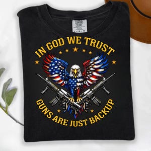 May include: Black t-shirt featuring an eagle with wings in red, white, and blue, holding two rifles. The text "IN GOD WE TRUST" and "GUNS ARE JUST BACKUP" are in yellow.