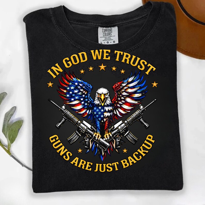May include: Black t-shirt featuring an eagle with wings in red, white, and blue, holding two rifles. The text "IN GOD WE TRUST" and "GUNS ARE JUST BACKUP" are in yellow.