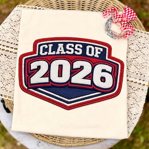 May include: Cream-colored t-shirt with a large embroidered patch that reads "CLASS OF 2026" in white, red, and blue. The patch is in a shield shape. A red and white checkered bow and bracelets are in the background.