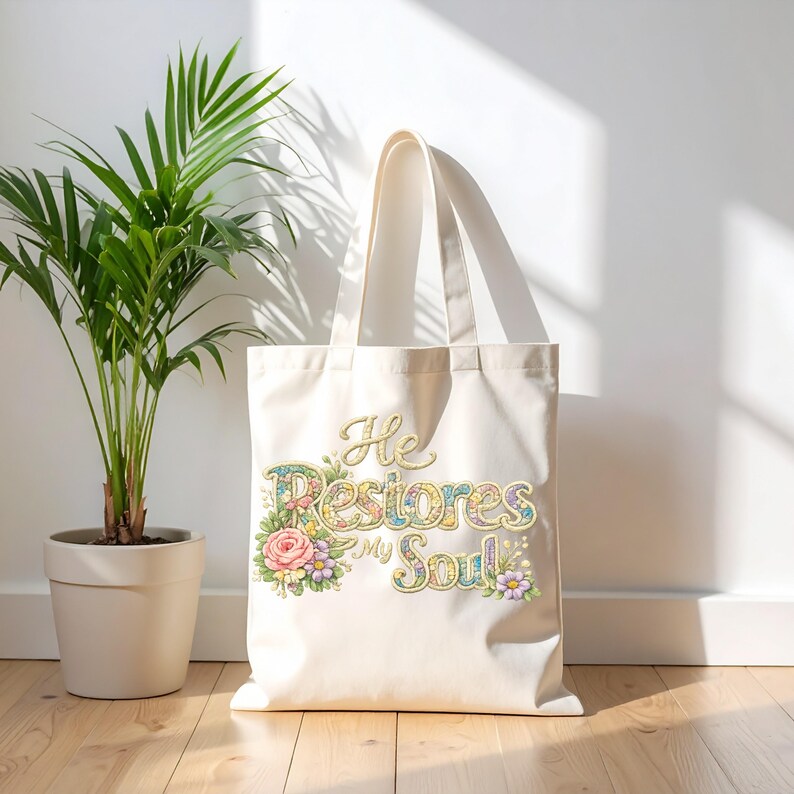 May include: Cream-colored tote bag with the text "He Restores My Soul" in a floral design. The bag has a simple design with two handles and is made of a sturdy fabric. A potted plant is to the left of the bag.