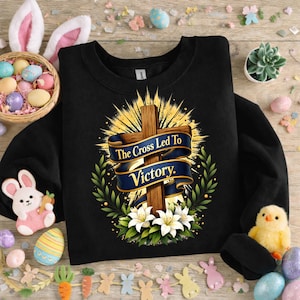 May include: Black crewneck sweatshirt with a graphic of a wooden cross, a blue banner with the text "The Cross Led To Victory," and white lilies. Easter-themed decorations surround the sweatshirt, including eggs and a bunny.