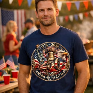 K&ouml;nnte beinhalten: Ein marineblaues T-Shirt mit einem patriotischen Design. Die Grafik enth&auml;lt den Text "1776-2026" und "250 AMERICAN HERITAGE" mit Bildern der Freiheitsstatue, der Liberty Bell und eines Adlers. Das T-Shirt wird von einer Person getragen.