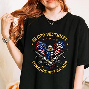 May include: Black t-shirt featuring a patriotic design with an eagle, American flag wings, and two rifles. The text reads "IN GOD WE TRUST" and "GUNS ARE JUST BACKUP".