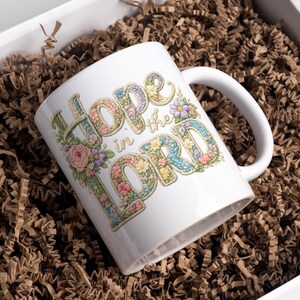 May include: White ceramic mug with the words "Hope in the Lord" in a floral design. The mug is inside a white box filled with brown paper. The letters are decorated with flowers in pastel colors.