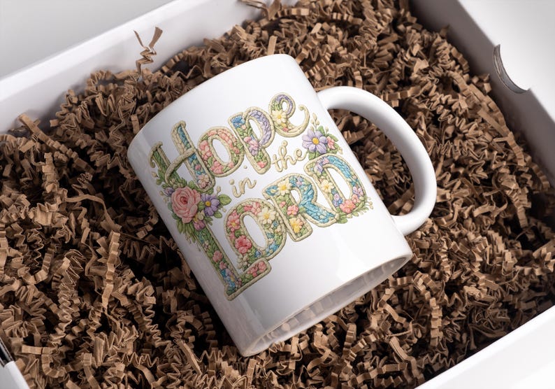 May include: White ceramic mug with the words "Hope in the Lord" in a floral design. The mug is inside a white box filled with brown paper. The letters are decorated with flowers in pastel colors.