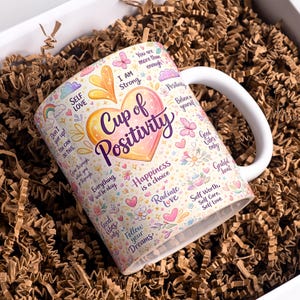 May include: White ceramic mug with a large heart design that reads "Cup of Positivity." The mug is decorated with inspirational phrases, flowers, butterflies, and hearts. The mug is packaged in a white box with brown crinkle paper.