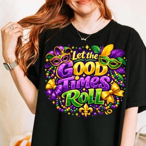 Op de afbeelding: Zwart T-shirt met een kleurrijk grafisch ontwerp. Het ontwerp toont de tekst "Let the GOOD Times Roll" in goud, paars en groen, samen met Mardi Gras-maskers, kralen en strikken. Onderaan staat een fleur-de-lis.