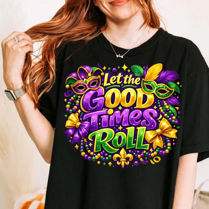 Op de afbeelding: Zwart T-shirt met een kleurrijk grafisch ontwerp. Het ontwerp toont de tekst "Let the GOOD Times Roll" in goud, paars en groen, samen met Mardi Gras-maskers, kralen en strikken. Onderaan staat een fleur-de-lis.