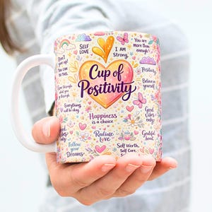 May include: White ceramic mug with a colorful design featuring the words "Cup of Positivity" in a heart shape, surrounded by inspirational phrases, flowers, and butterflies. The mug has a white handle and is held by a person.