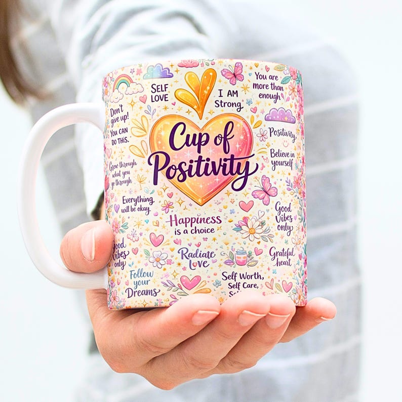 May include: White ceramic mug with a colorful design featuring the words "Cup of Positivity" in a heart shape, surrounded by inspirational phrases, flowers, and butterflies. The mug has a white handle and is held by a person.