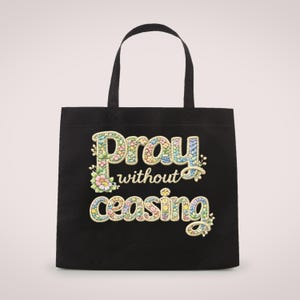 May include: Black tote bag with the words "Pray without ceasing" in colorful, floral-style lettering. The bag has two black handles and a simple design, suitable for carrying various items.