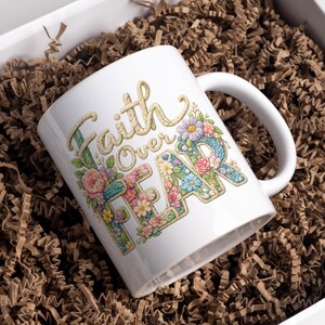 May include: White ceramic mug with the words "Faith Over Fear" in a floral design. The mug is decorated with colorful flowers and is presented in a white gift box with brown paper filler.