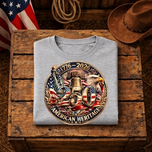 K&ouml;nnte beinhalten: Ein graues T-Shirt mit einem kreisf&ouml;rmigen Grafikdesign, das das 250-j&auml;hrige Jubil&auml;um des amerikanischen Erbes feiert. Das Design zeigt die Freiheitsstatue, die Liberty Bell, einen Adler und die amerikanische Flagge. Der Text "1776-2026" und "American Heritage" sind ebenfalls enthalten.