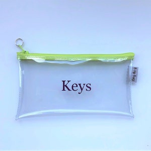 May include: A clear plastic zippered pouch with a lime green zipper and trim. The pouch has the word "Keys" printed on it in purple and a small white tag with the text "B Hip Bags" on the right side.