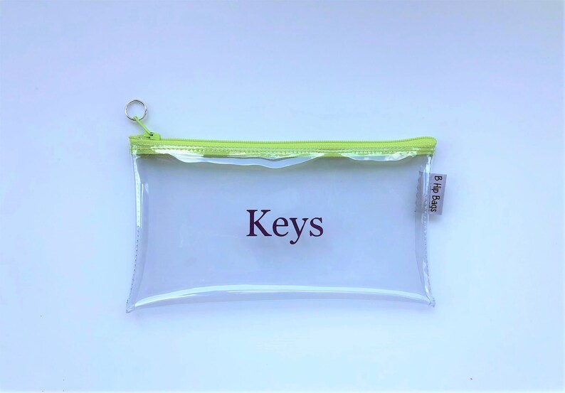 May include: A clear plastic zippered pouch with a lime green zipper and trim. The pouch has the word "Keys" printed on it in purple and a small white tag with the text "B Hip Bags" on the right side.