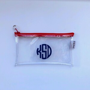 May include: Clear plastic zippered pouch with a red zipper and a navy blue monogram "KSD" in a circle. The pouch has a small silver ring attached to the zipper.