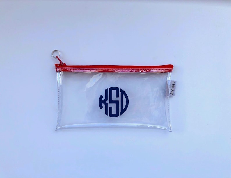 May include: Clear plastic zippered pouch with a red zipper and a navy blue monogram "KSD" in a circle. The pouch has a small silver ring attached to the zipper.