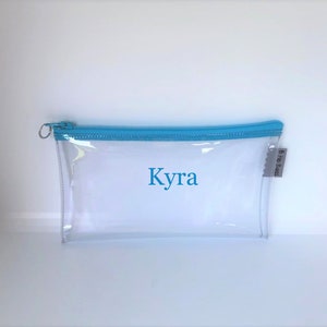 May include: Clear plastic pencil case with a light blue zipper and the name "Kyra" printed in blue on the front. The case has a small tag with the text "B Ho Bags" on the side.
