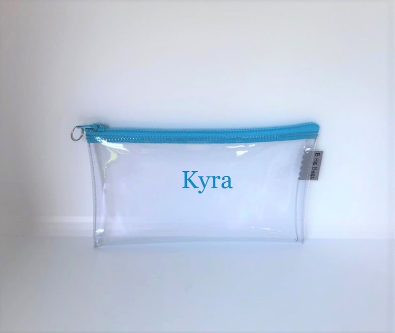 May include: Clear plastic pencil case with a light blue zipper and the name "Kyra" printed in blue on the front. The case has a small tag with the text "B Ho Bags" on the side.