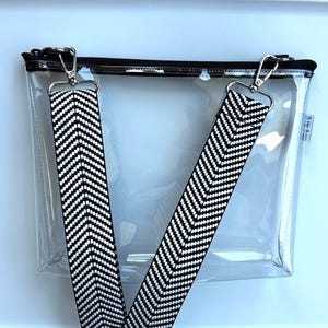 May include: A clear, rectangular crossbody bag with a black zipper and silver-tone hardware. The bag features two detachable straps with a black and white chevron pattern. The bag is designed for carrying essentials.