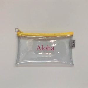 May include: Clear plastic pencil case with a yellow zipper and a pink "Aloha" inscription. The case has a small white label with the text "B.Hip Bags".