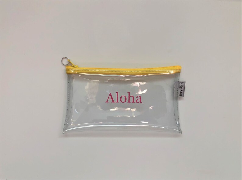 May include: Clear plastic pencil case with a yellow zipper and a pink "Aloha" inscription. The case has a small white label with the text "B.Hip Bags".