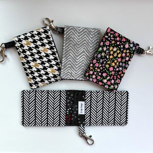 May include: A collection of fabric wallets and keychains in various patterns. Designs include a black and white houndstooth, gray chevron, and a floral print on black. One wallet is open, displaying a black and white herringbone pattern.