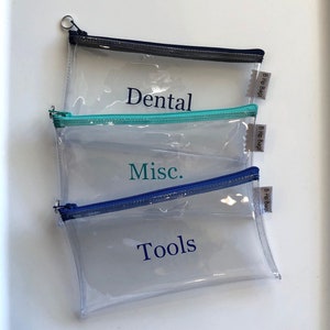 May include: Three clear plastic zippered pouches with blue, teal, and gray zippers. The pouches are labeled "Dental", "Misc.", and "Tools" in blue text. Each pouch has a small white label with the text "B Hip Bags" on it.