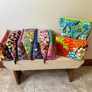 May include: A collection of colorful, patterned cosmetic bags and pouches. The bags feature floral, paisley, and abstract designs in various colors, including blue, orange, pink, and yellow. They are displayed on a small, white bench with wooden legs.