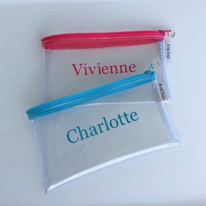 May include: Two clear plastic zippered pouches with personalized names. The top pouch has a pink zipper and the name "Vivienne" printed in pink. The bottom pouch has a blue zipper and the name "Charlotte" printed in teal. Both pouches have a white label with the text "B Hip Bags".