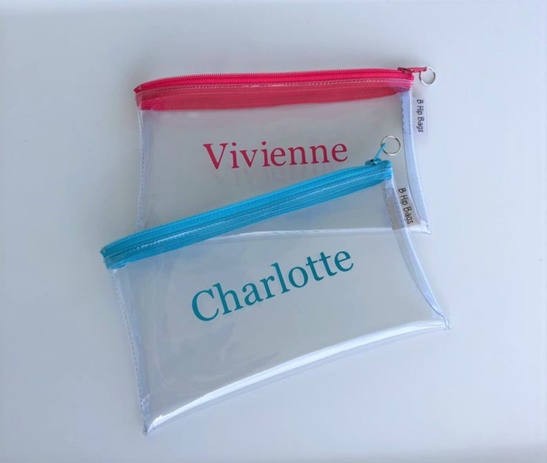 May include: Two clear plastic zippered pouches with personalized names. The top pouch has a pink zipper and the name "Vivienne" printed in pink. The bottom pouch has a blue zipper and the name "Charlotte" printed in teal. Both pouches have a white label with the text "B Hip Bags".