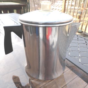 May include: A silver percolator coffee pot with a black handle. The pot has a shiny, reflective surface.