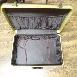 May include: An open vintage suitcase with a cream-colored exterior frame and a dark brown interior lining. The lid features a gathered fabric pocket, while the base includes thin brown fabric straps for securing items inside the luggage.