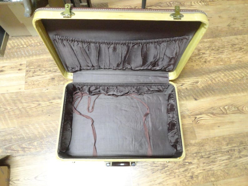May include: An open vintage suitcase with a cream-colored exterior frame and a dark brown interior lining. The lid features a gathered fabric pocket, while the base includes thin brown fabric straps for securing items inside the luggage.