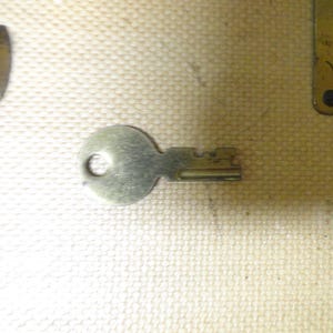 May include: A small, silver-toned metal key with a circular head and a notched blade. The key features a simple design with a hole in the center of the head for a keyring. It is positioned horizontally, showing a metallic finish and distinct teeth.