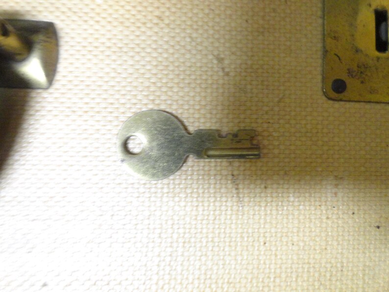 May include: A small, silver-toned metal key with a circular head and a notched blade. The key features a simple design with a hole in the center of the head for a keyring. It is positioned horizontally, showing a metallic finish and distinct teeth.
