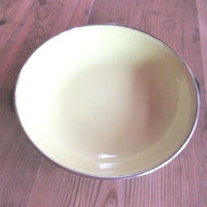 May include: A round, pale yellow bowl with a silver rim and two small handles. The bowl is empty and sits on a wooden surface. The bowl's interior is a solid, light yellow color.