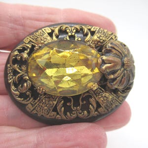 May include: An oval brooch featuring a large, faceted yellow glass stone set against a dark brown base. The stone is surrounded by intricate, raised gold-tone metal filigree work and a floral-shaped metal accent on the right side.