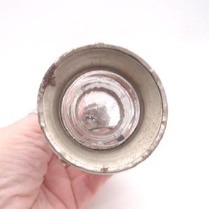 Pu&ograve; includere: Un tappo per bottiglia vintage con base in vetro trasparente e parte superiore in metallo color argento ossidato. Il tappo &egrave; tenuto in mano su sfondo bianco. Il metallo mostra segni di et&agrave; e usura.